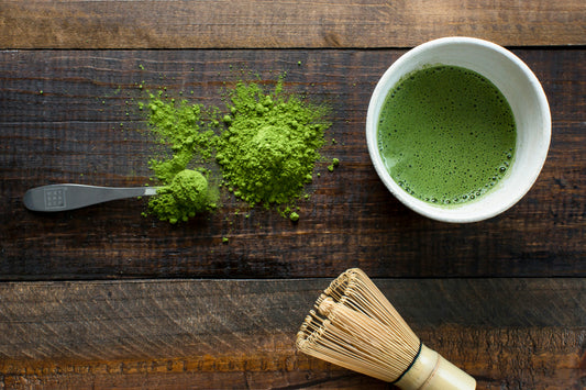 A bowl of prepared matcha tea on a wooden surface, with a heap of matcha powder, a spoon, and a bamboo whisk, showcasing the tea-making process