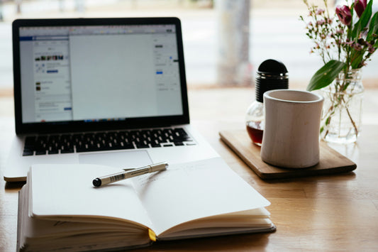 A modern workspace featuring an open laptop displaying social media pages, a notepad, pen, and a ceramic mug beside a vase with fresh flowers on a wooden table