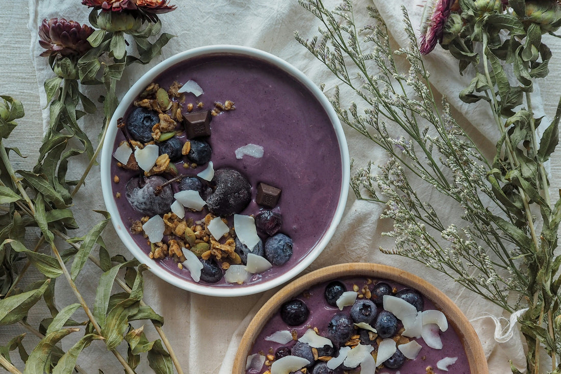 Açaí smoothie bowl topped with blueberries, granola, coconut flakes, and chocolate pieces, surrounded by dried flowers