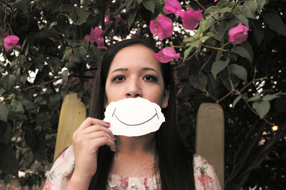 Woman holding a paper with a drawn smile covering her mouth, symbolizing masking emotions